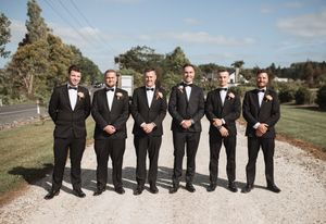 Groomsmen in black tuxedos line up for a formal photo on a gravel path.