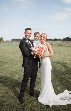 A wedding couple shares a joyful moment together in a grassy field with their bouquet of pink and white flowers.