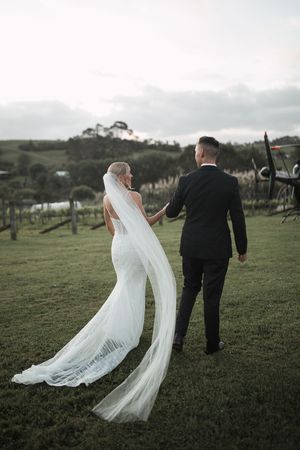 A bride's veil flows dramatically in the wind as couple walks across green hillside.