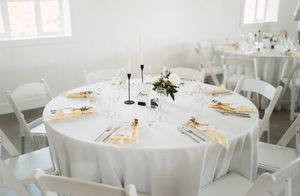 Elegant white wedding reception table setting with gold accents and black candlesticks in a bright room.