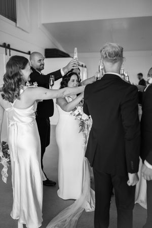 Wedding party in black suits and cream dresses posing outdoors against white building.