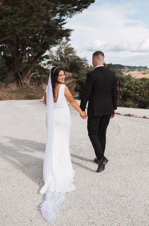 Bride in sleek white dress and groom walking hand in hand on scenic mountain path.