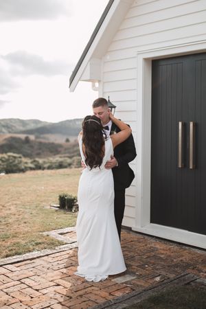 A couple in wedding attire shares a tender moment outside a white building with black doors.