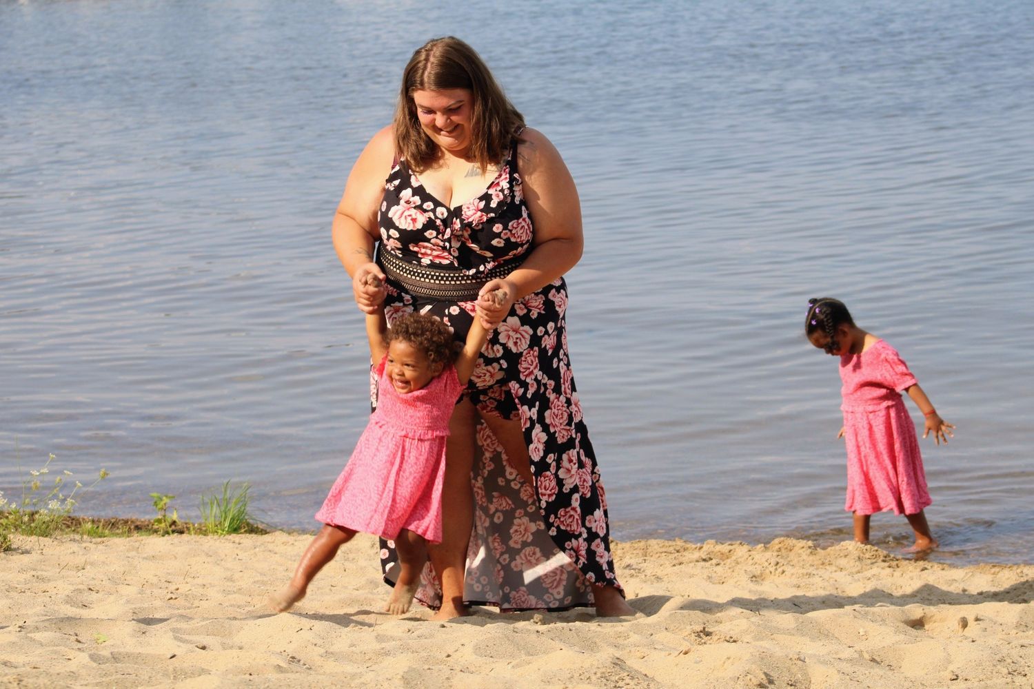 Woman in floral dress playing with two young girls in pink dresses on a sandy beach by the water.