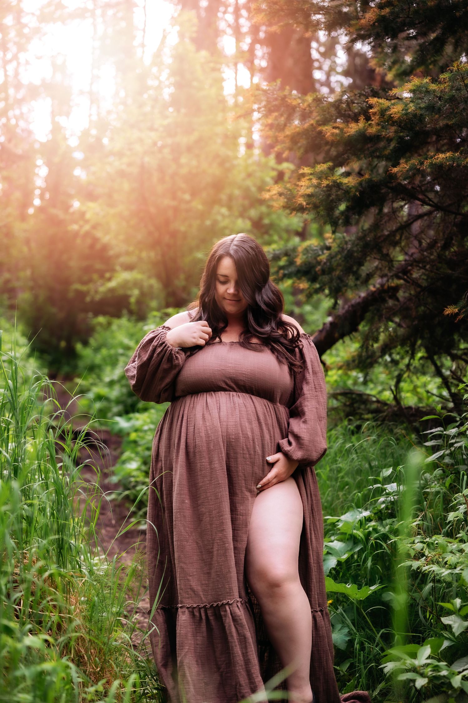Maternity photo of woman in flowing mauve dress standing in sunlit forest.