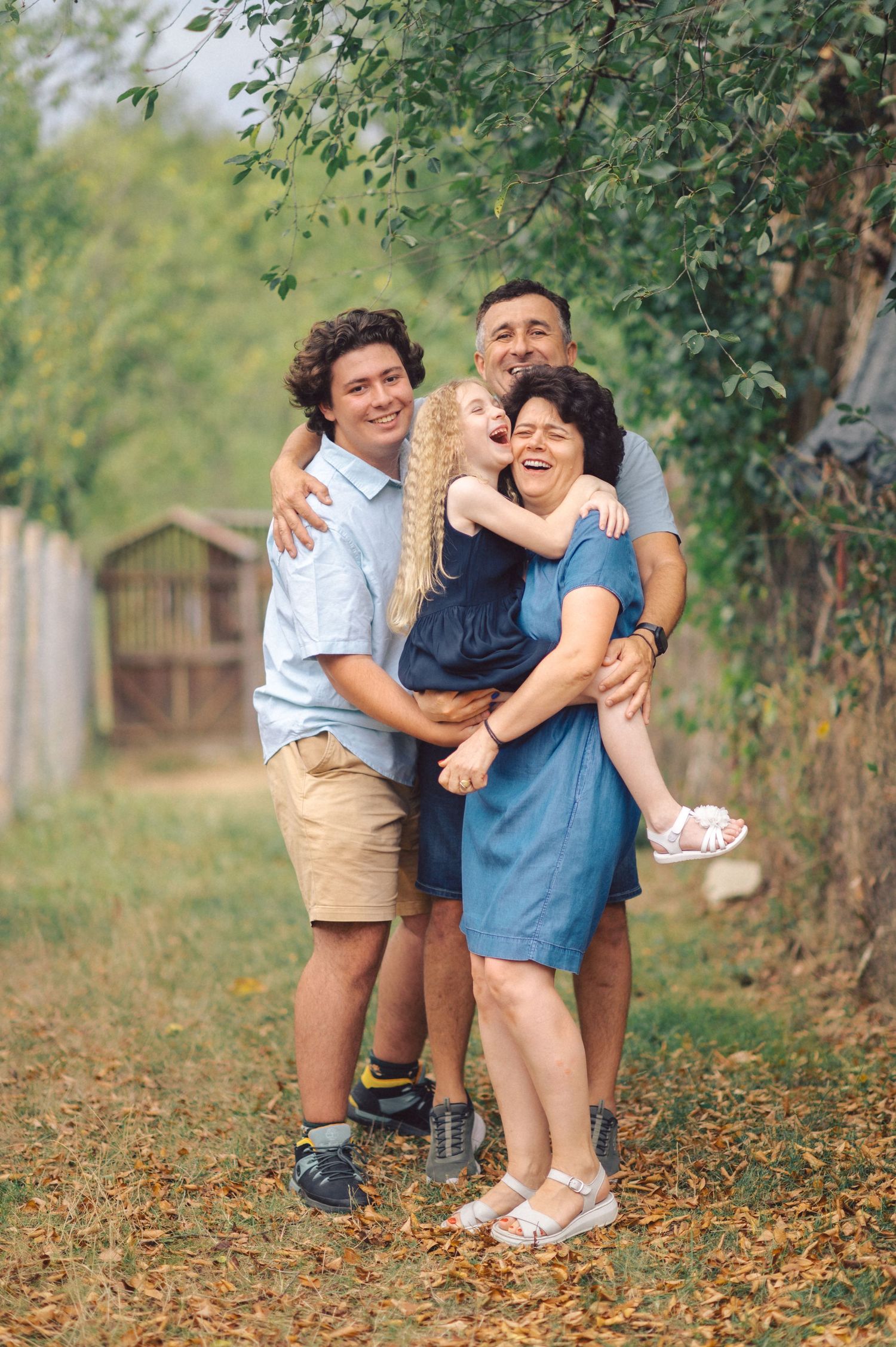 A family shares a warm embrace while standing on a leaf-covered path near a wooden shed in a garden setting.