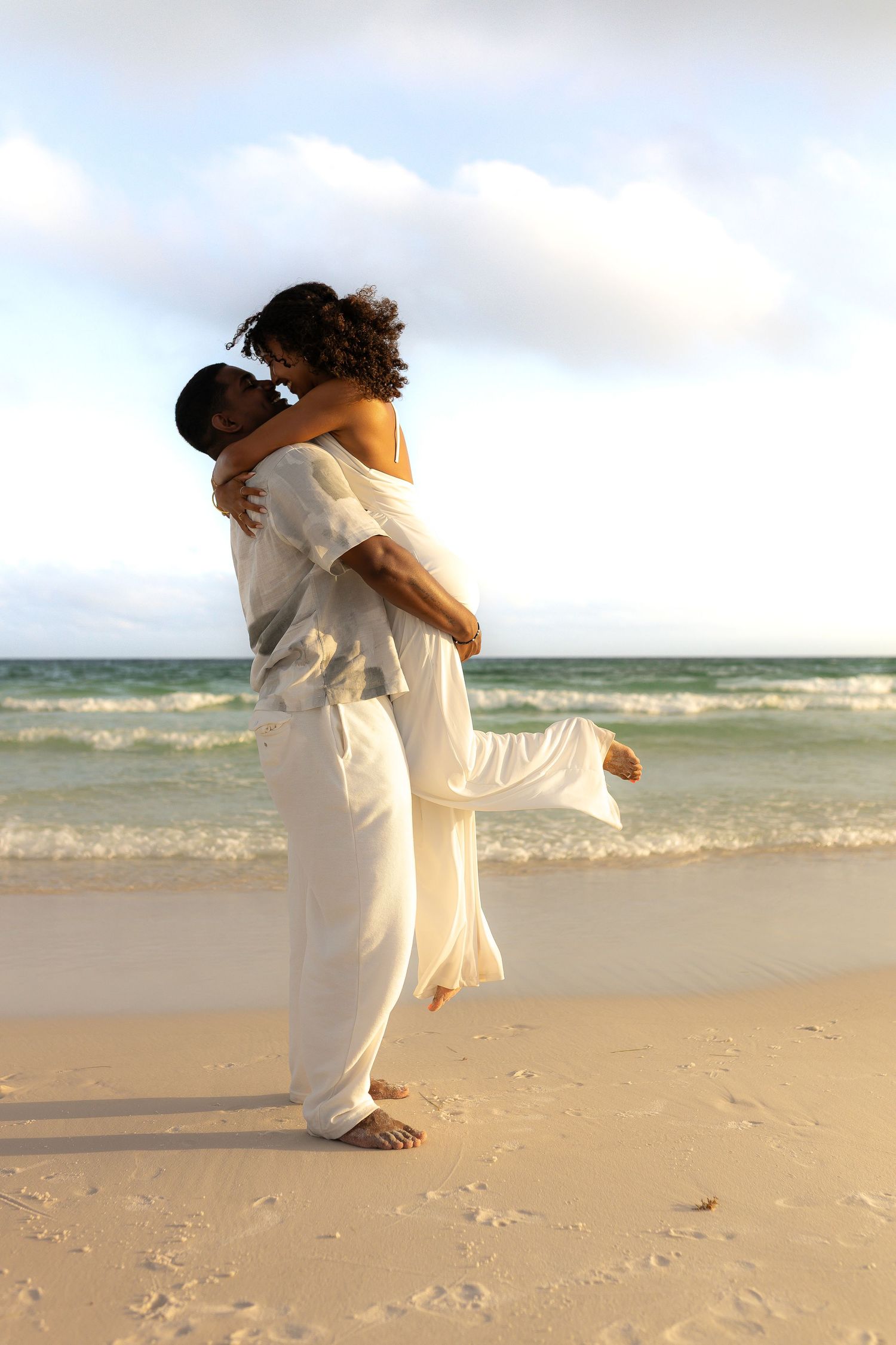 Romantic embrace on beach with flowing white outfits at sunset.