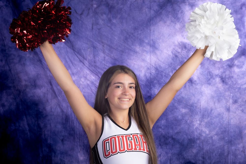 Young cheerleader in Cougars uniform holds red and white pom-poms overhead, smiling against purple backdrop.