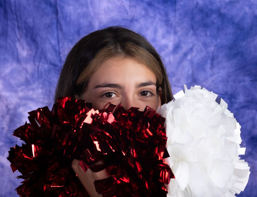 Cheerleader playfully hides face behind red and white pom-poms against blue backdrop.