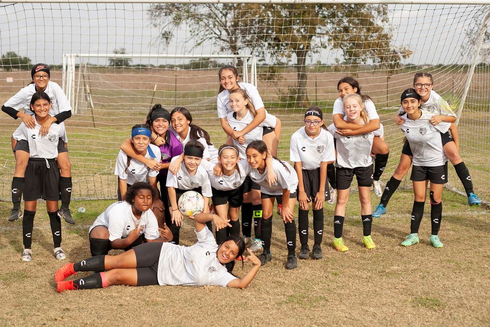 Girls soccer team posing together in black and white uniforms in front of a goal on a grass field.