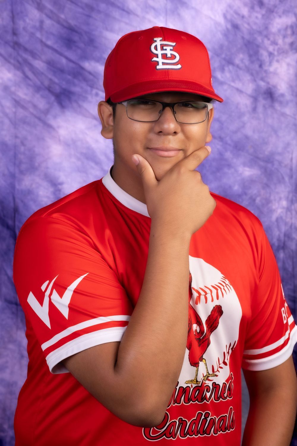 Young baseball player in red Cardinals uniform and cap poses thoughtfully with hand on chin.