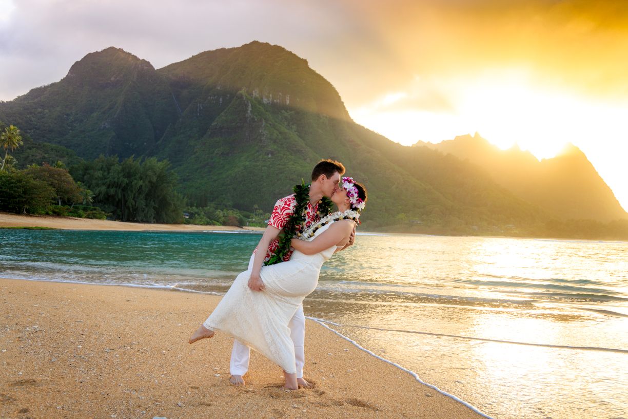 A couple embraces during sunset on a tropical beach with mountains and golden light in Hanalei Bay, Hawaii.