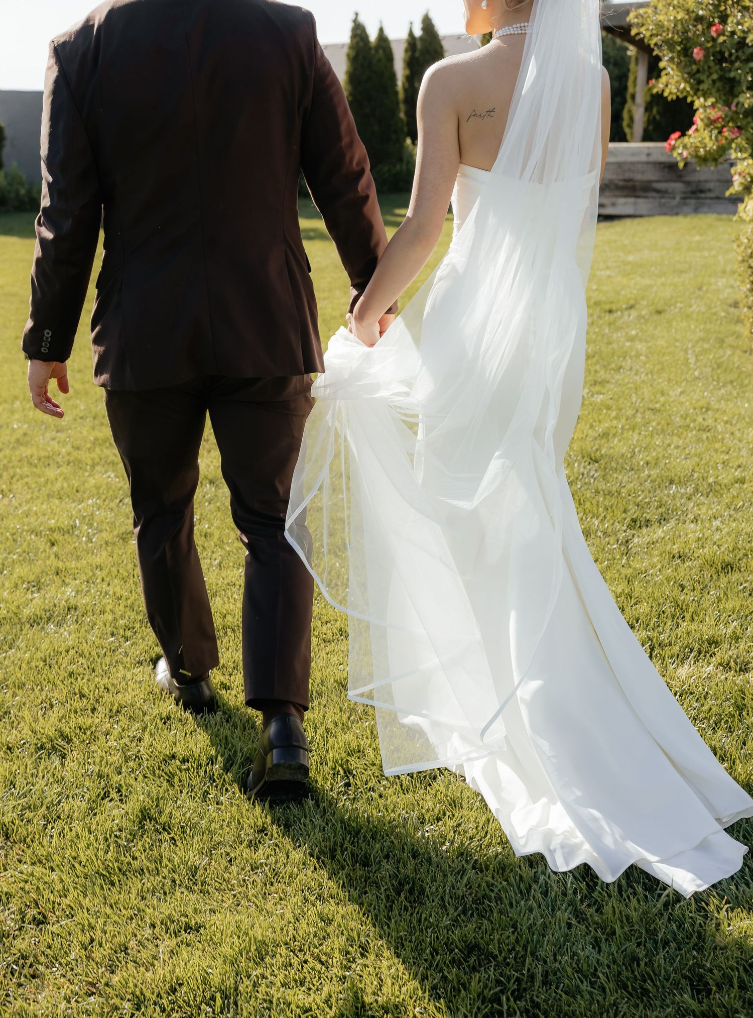 Bride and groom walking hand in hand on green lawn, bride in flowing white gown with elegant train.