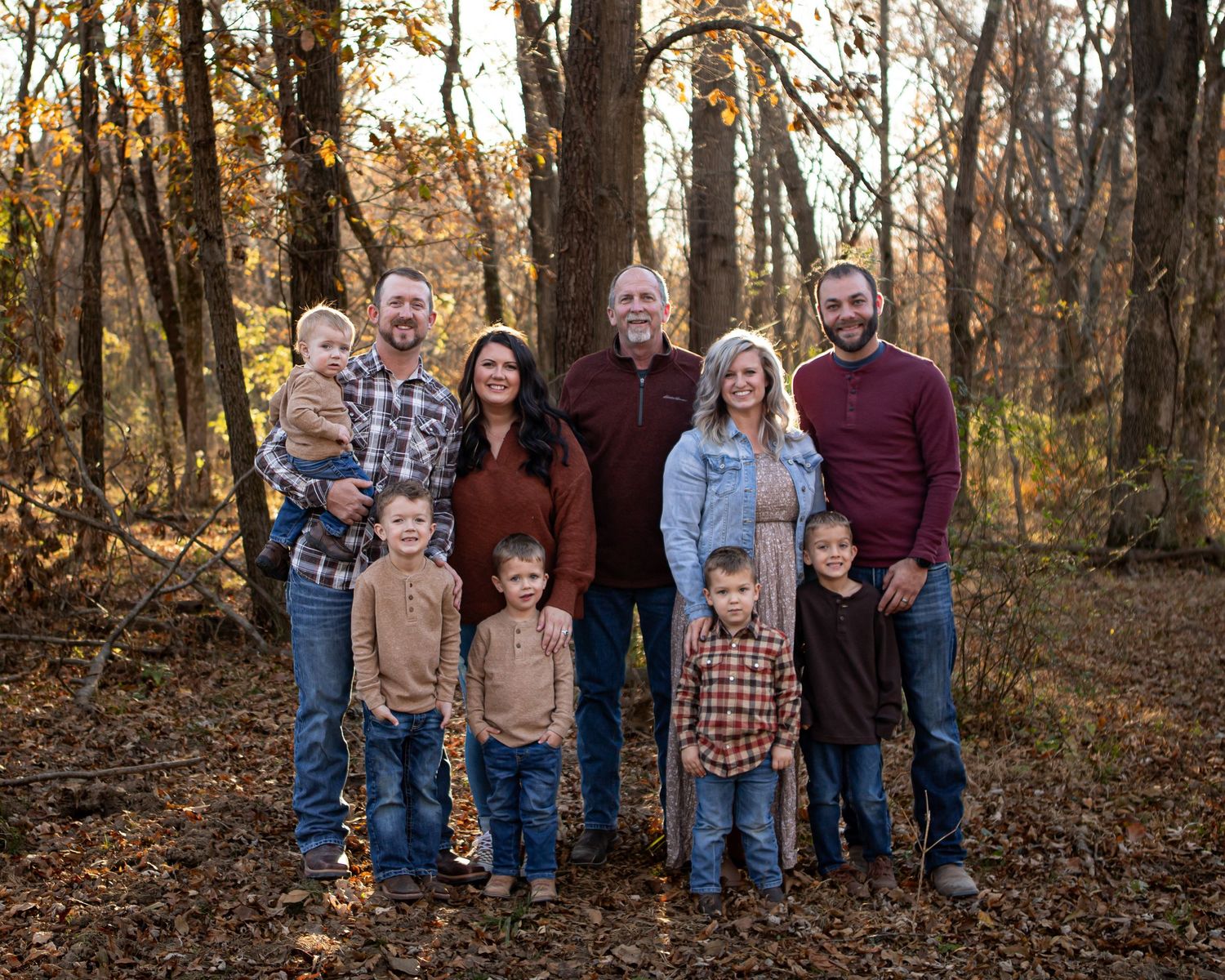 Extended family poses together for autumn portrait in wooded setting with fallen leaves.