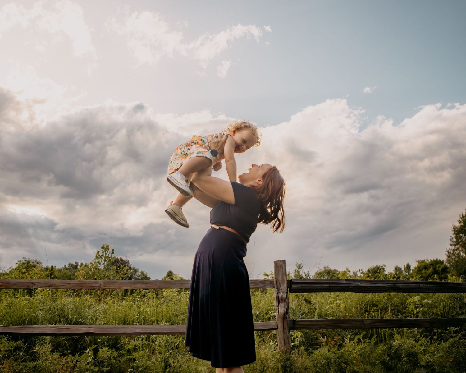 Woman in black dress poses against fence with dramatic cloudy sky background.