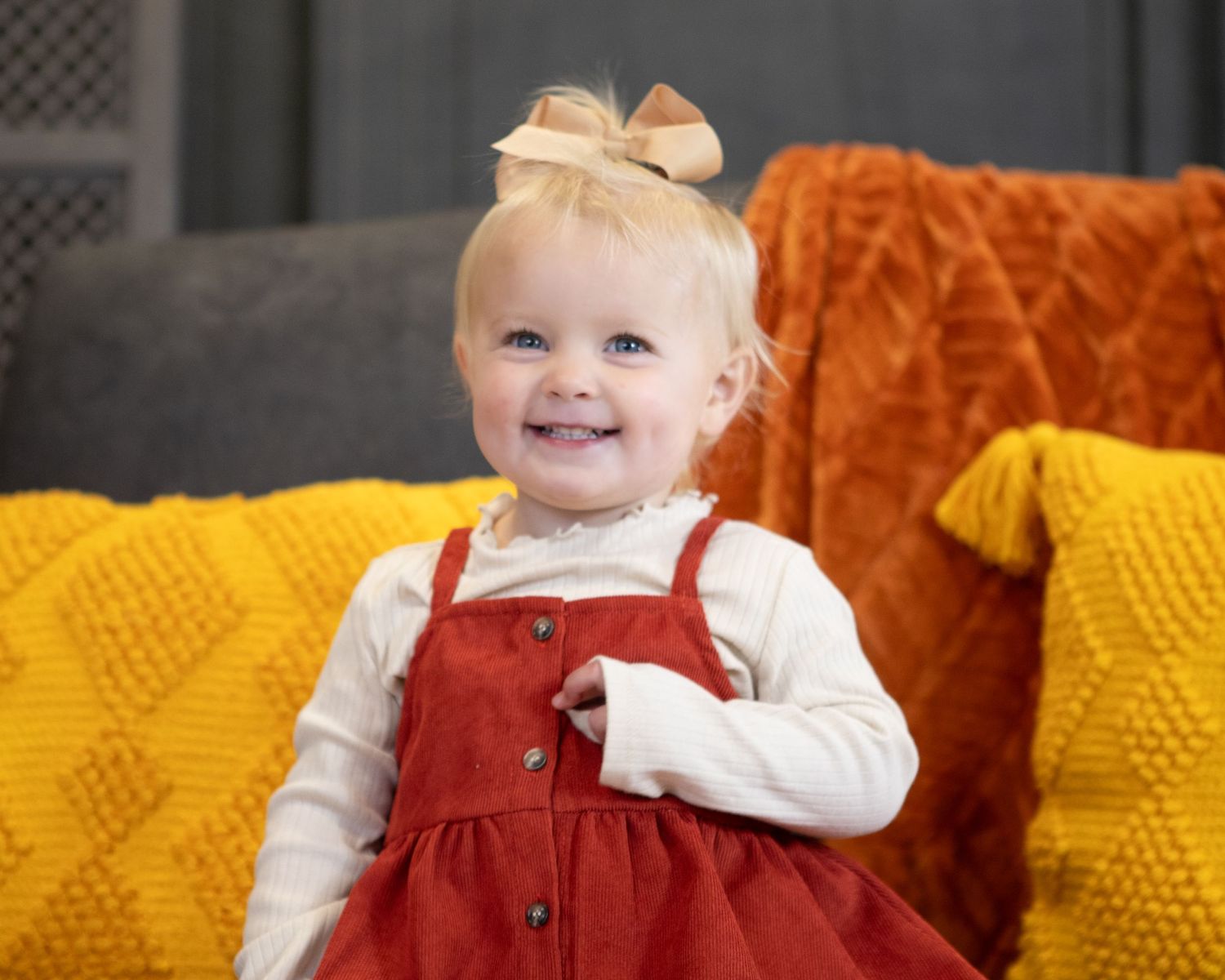 Young child wearing a red corduroy jumper dress sits against orange and yellow pillows.