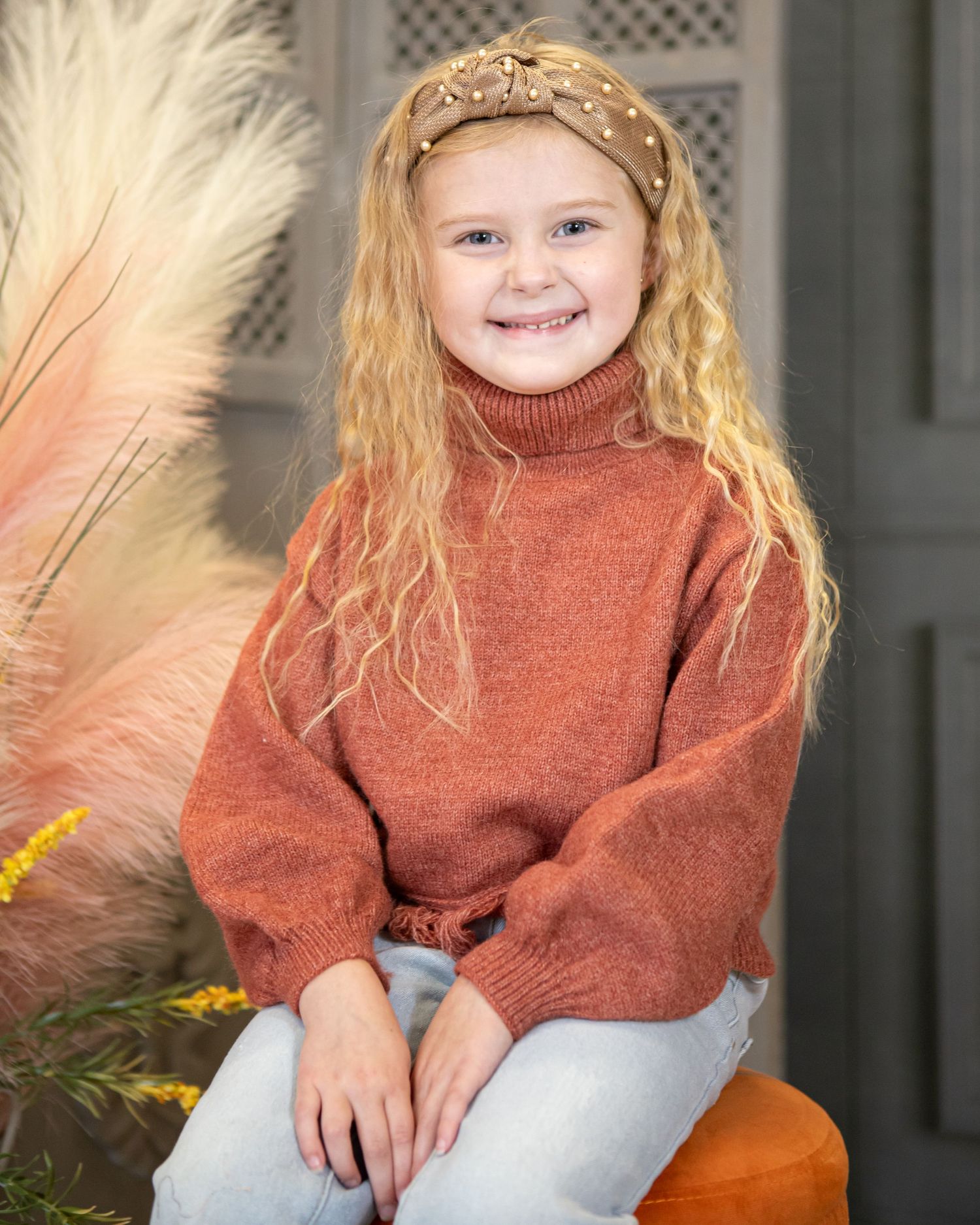 Child in rust colored sweater sits on orange pumpkin for autumn photo.