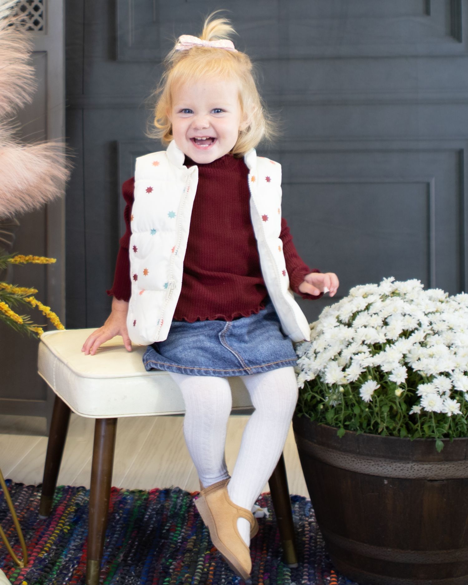 Child in burgundy top and white vest sits by white flowers outdoors.