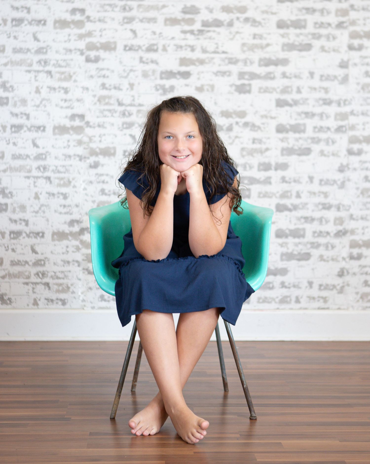 Person in navy dress sits curled up on turquoise chair against brick wall.