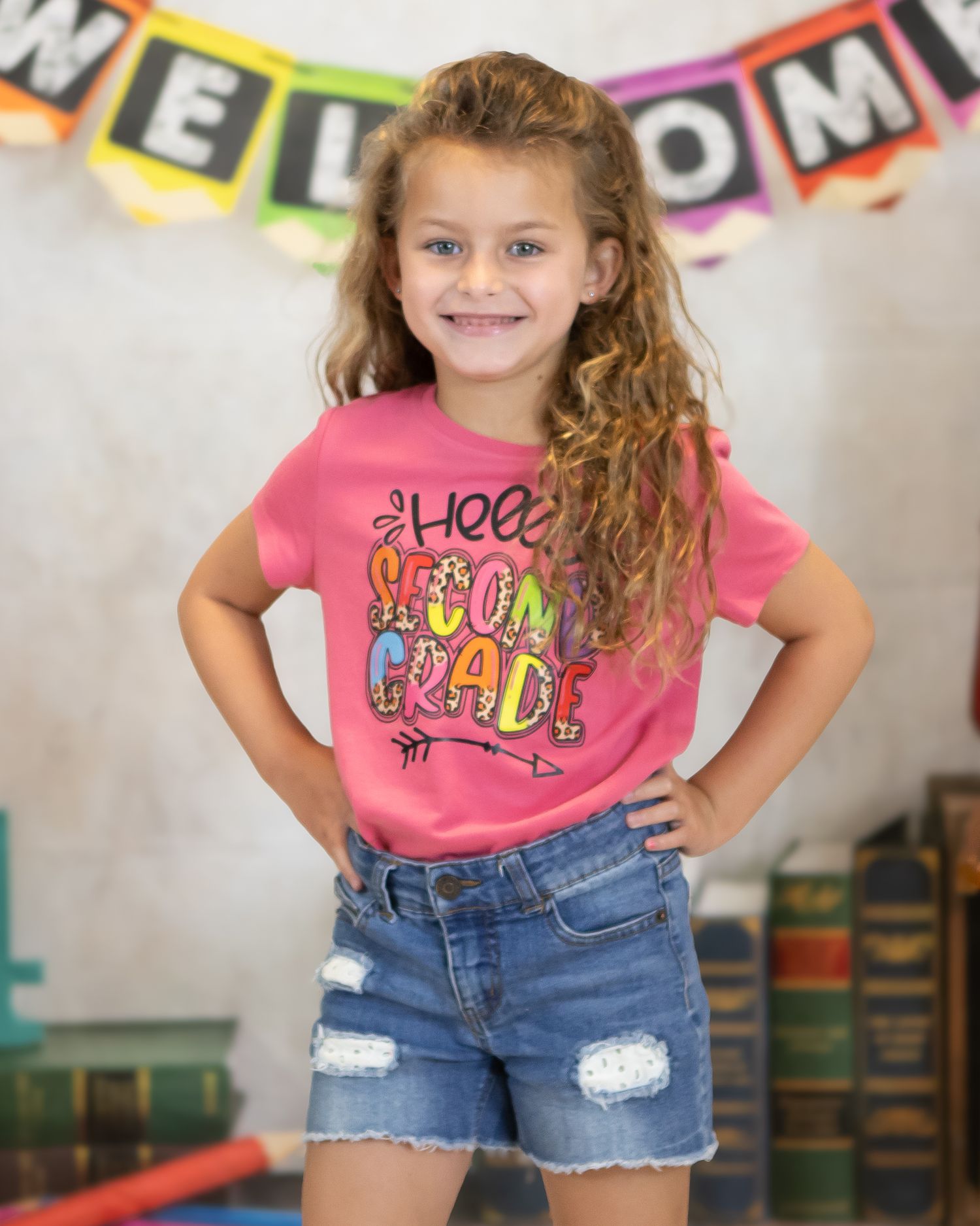 Child wearing pink graphic t-shirt and denim shorts poses under welcome banner.