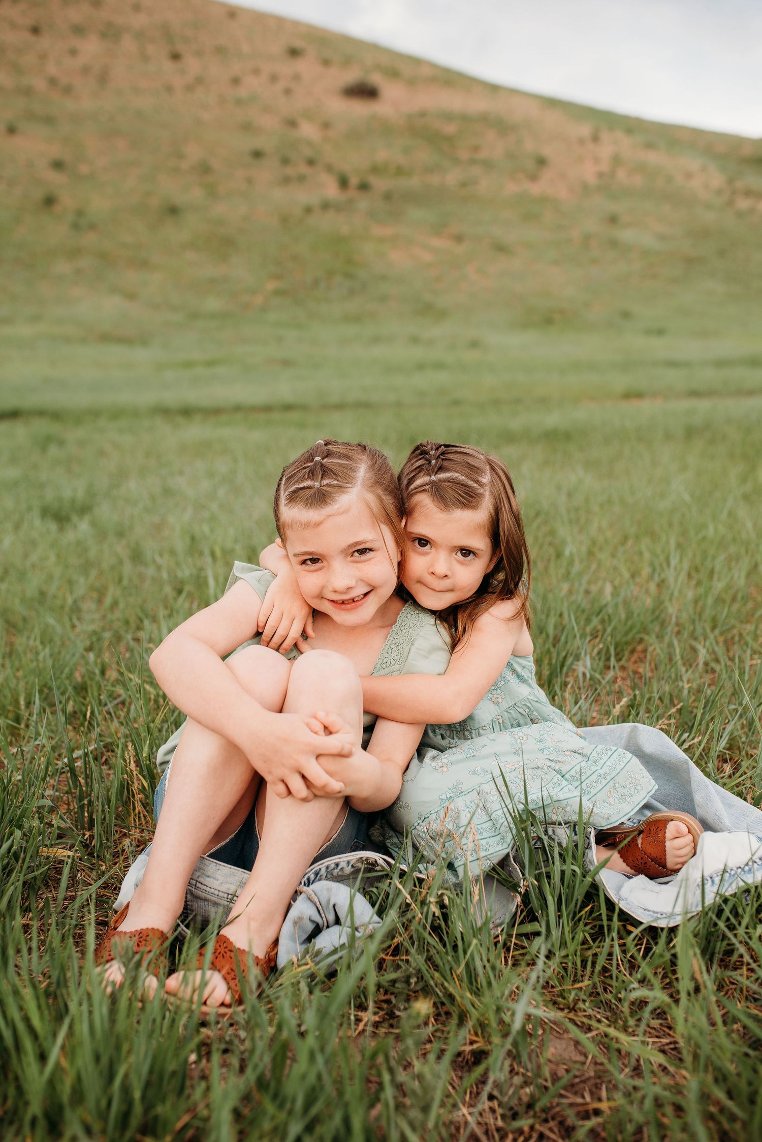 Two children embracing while sitting in tall grass on hillside during sunset.