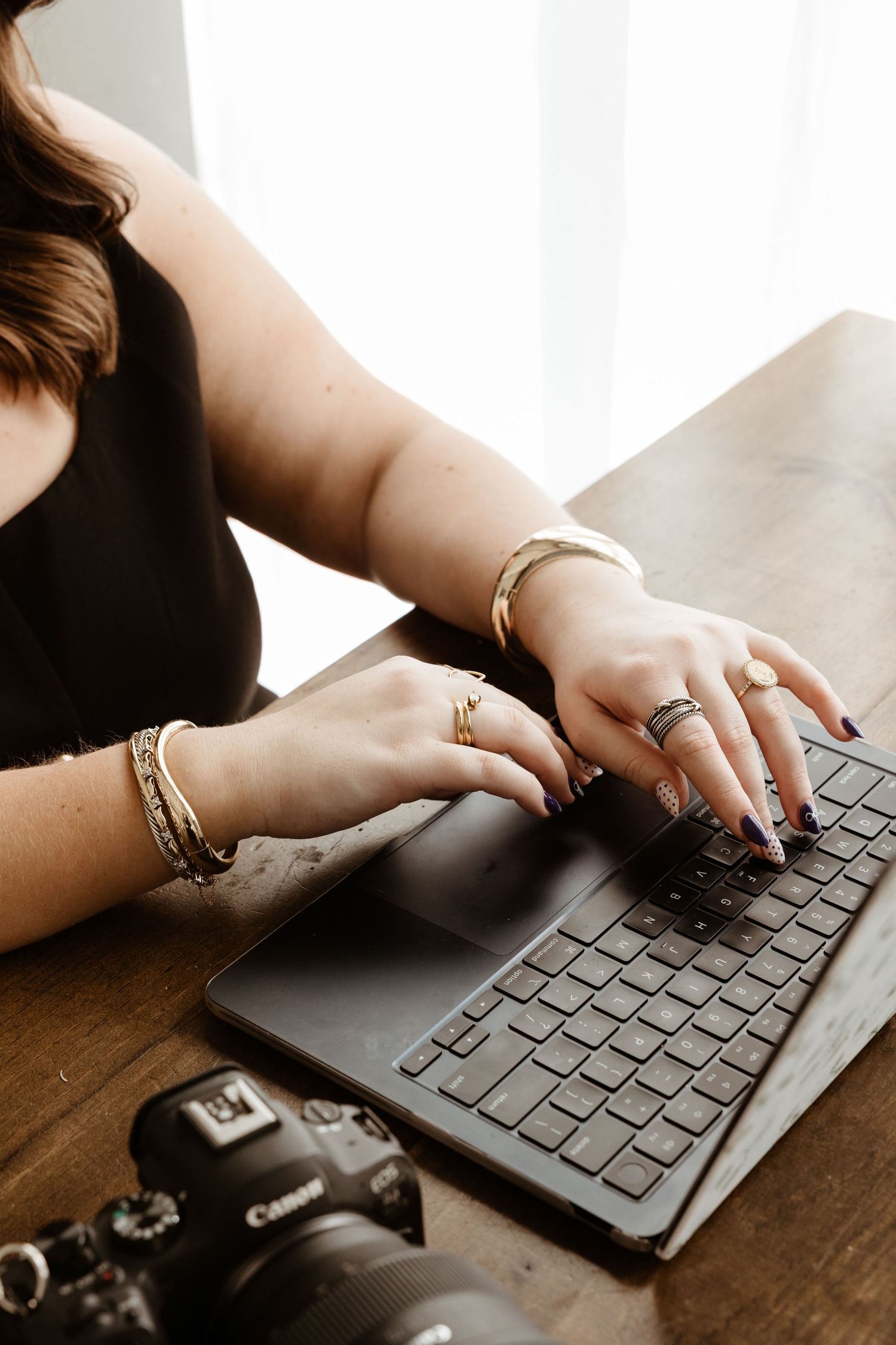 Woman with gold bracelets and rings typing on laptop beside Canon camera on wood desk.