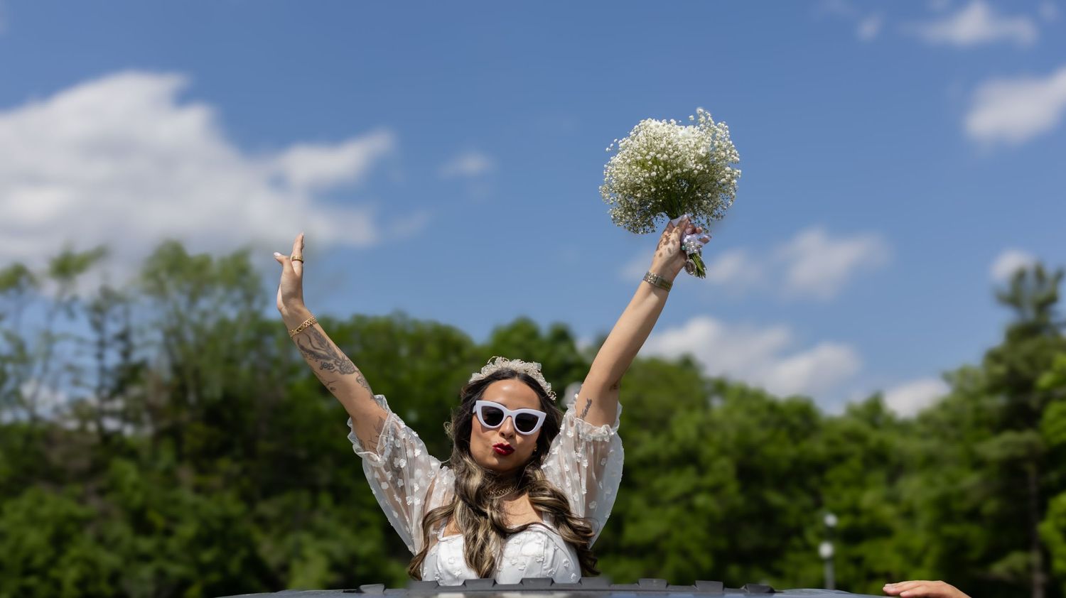Joyful bride in sunglasses holding bouquet against blue summer sky.