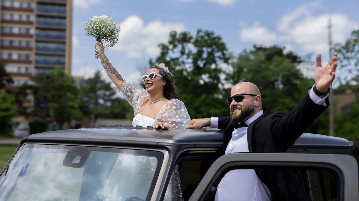 Newlyweds celebrating through sunroof of car with arms raised in celebration.