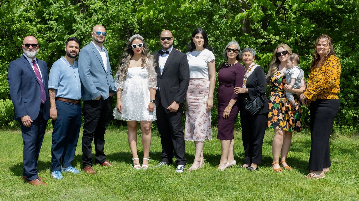 Wedding party group photo with guests lined up outdoors in summer garden setting.