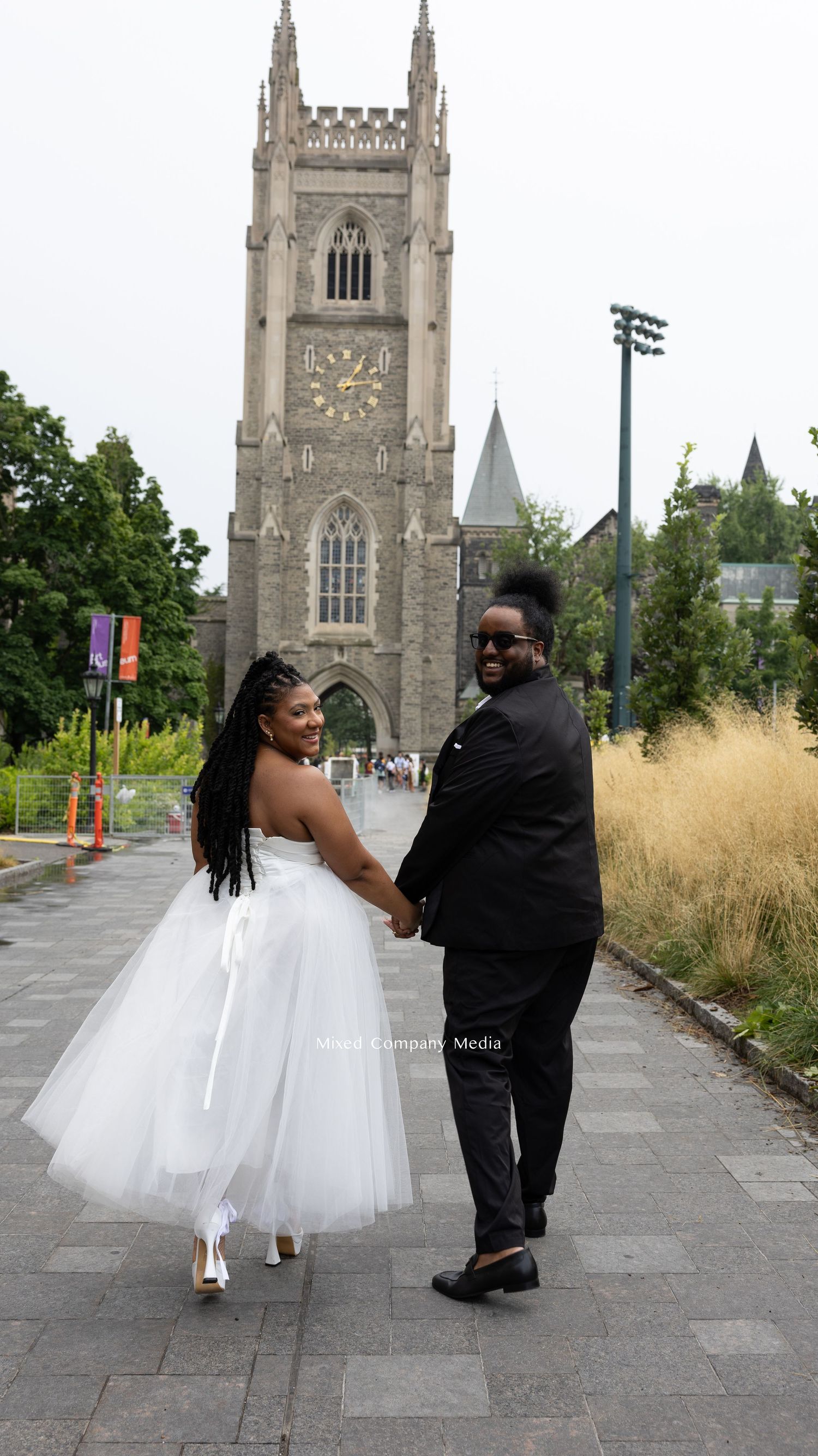 Wedding couple walking hand in hand in front of Gothic cathedral with clock tower.