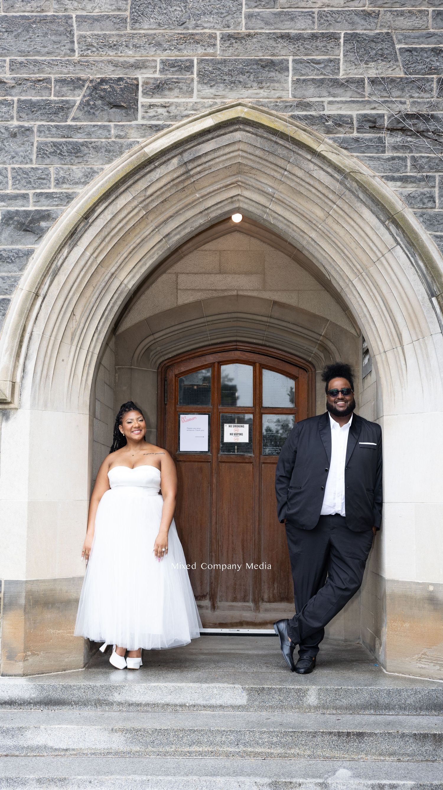 Wedding couple posing under stone archway entrance of historic building.