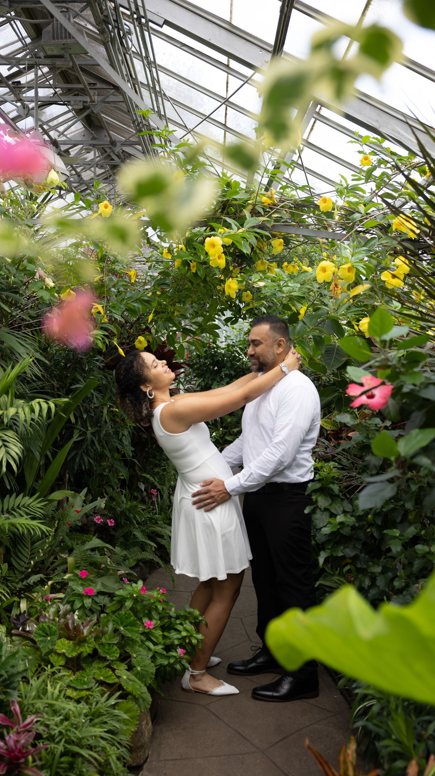 Intimate greenhouse photoshoot with couple surrounded by blooming yellow and pink flowers.