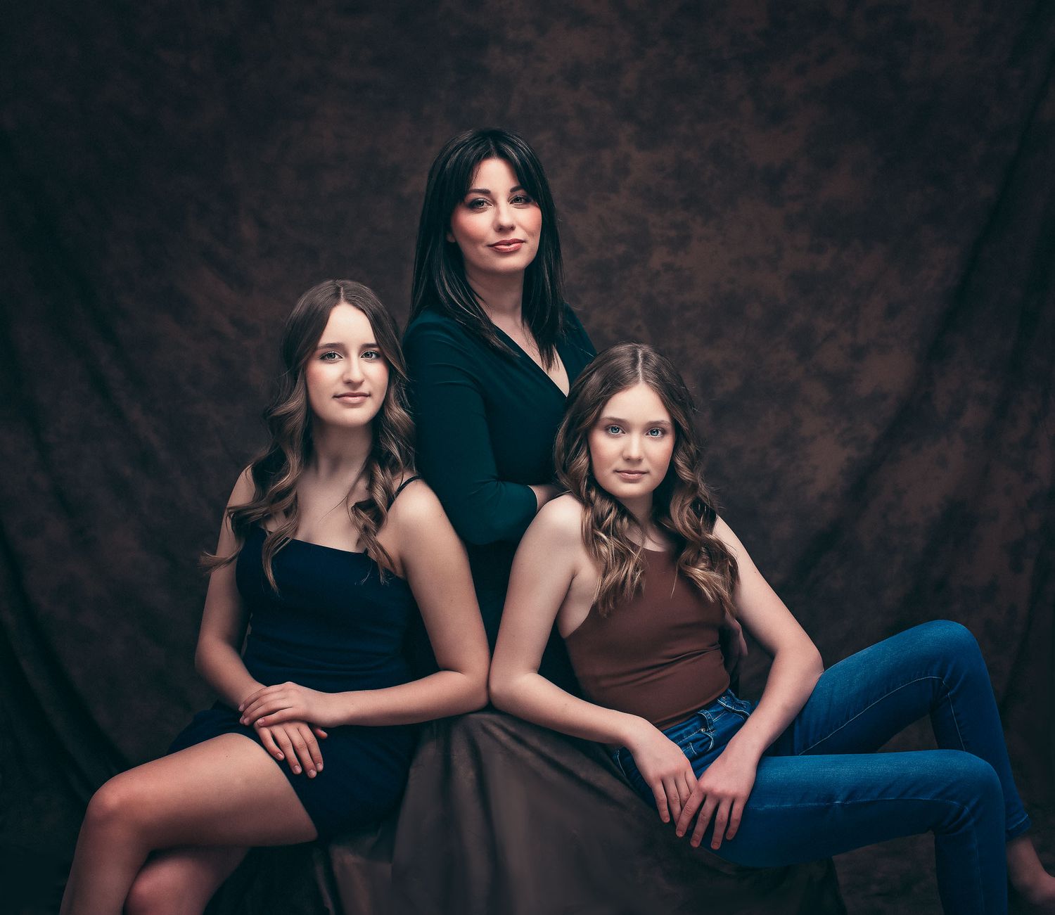 Three women in elegant attire pose together against a dark backdrop for a portrait photo.