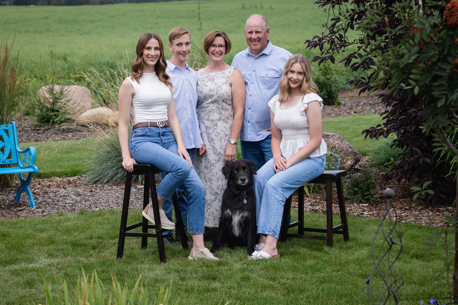 A family sits together on wooden stools in a lush green garden setting.