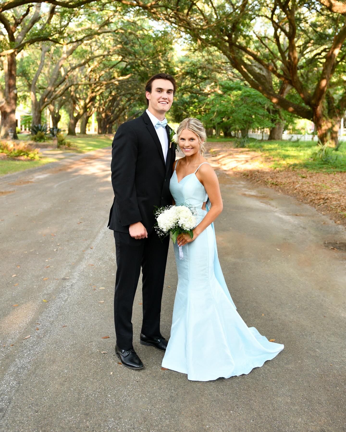 Formal couple poses on tree-lined path, bride in light blue dress with white bouquet.