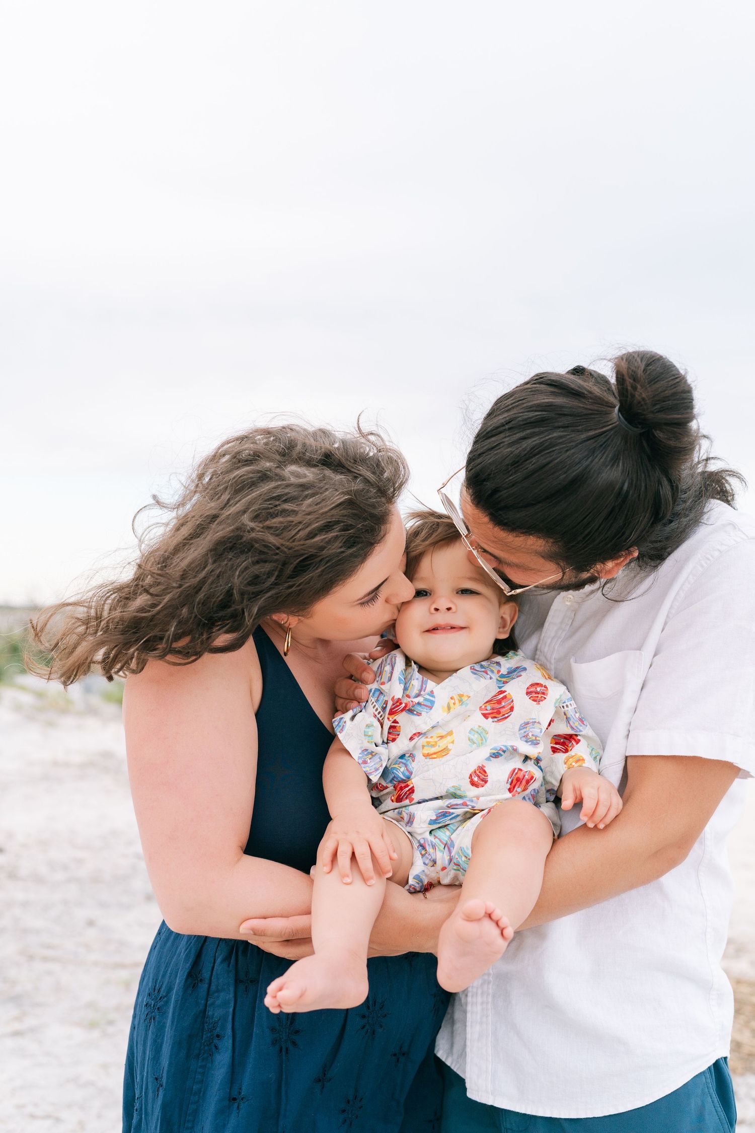 Parents kissing their smiling toddler’s cheeks during a beach family photography session.