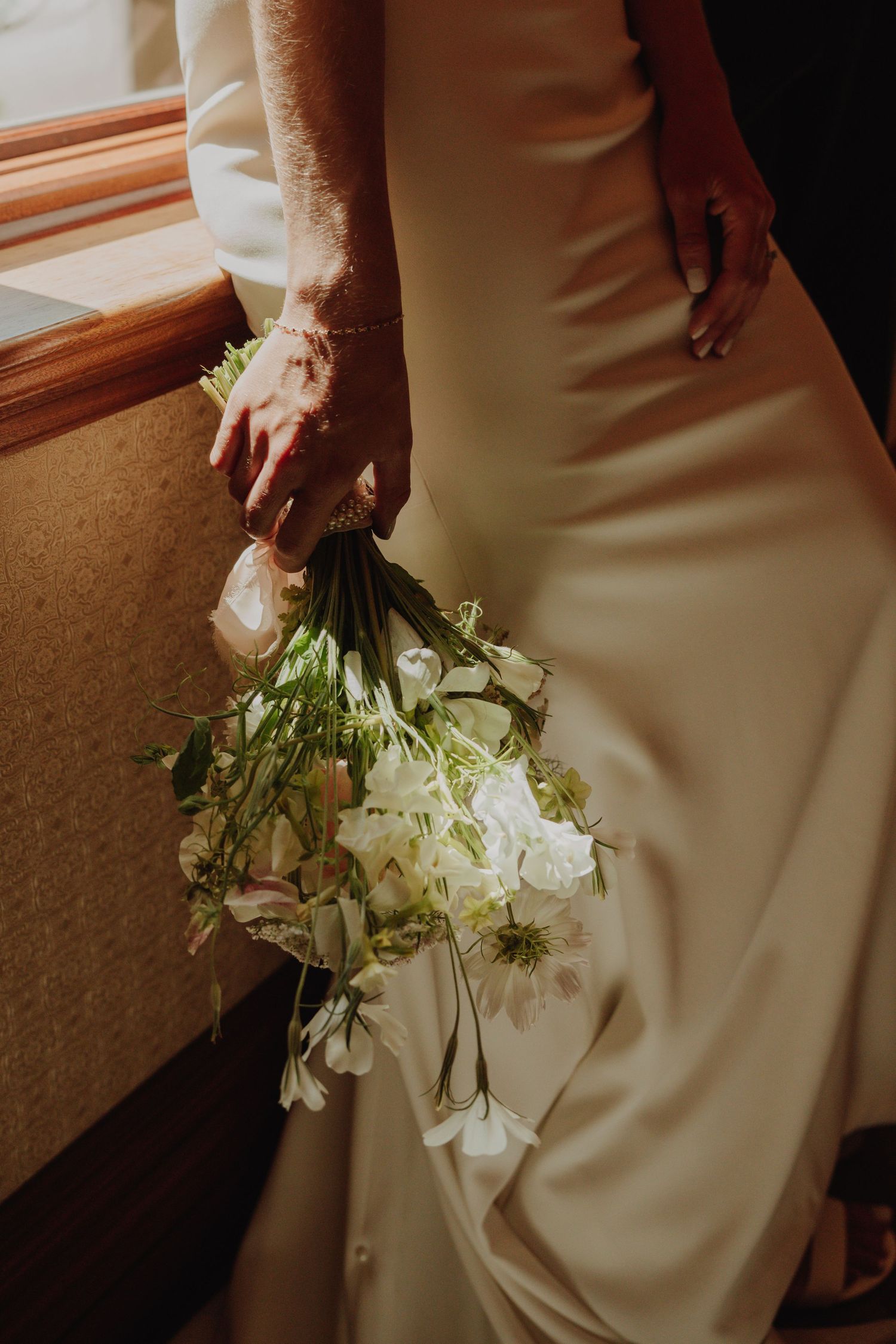 Bride in white satin gown holding an elegant bouquet of white roses and greenery beside a sunlit window.