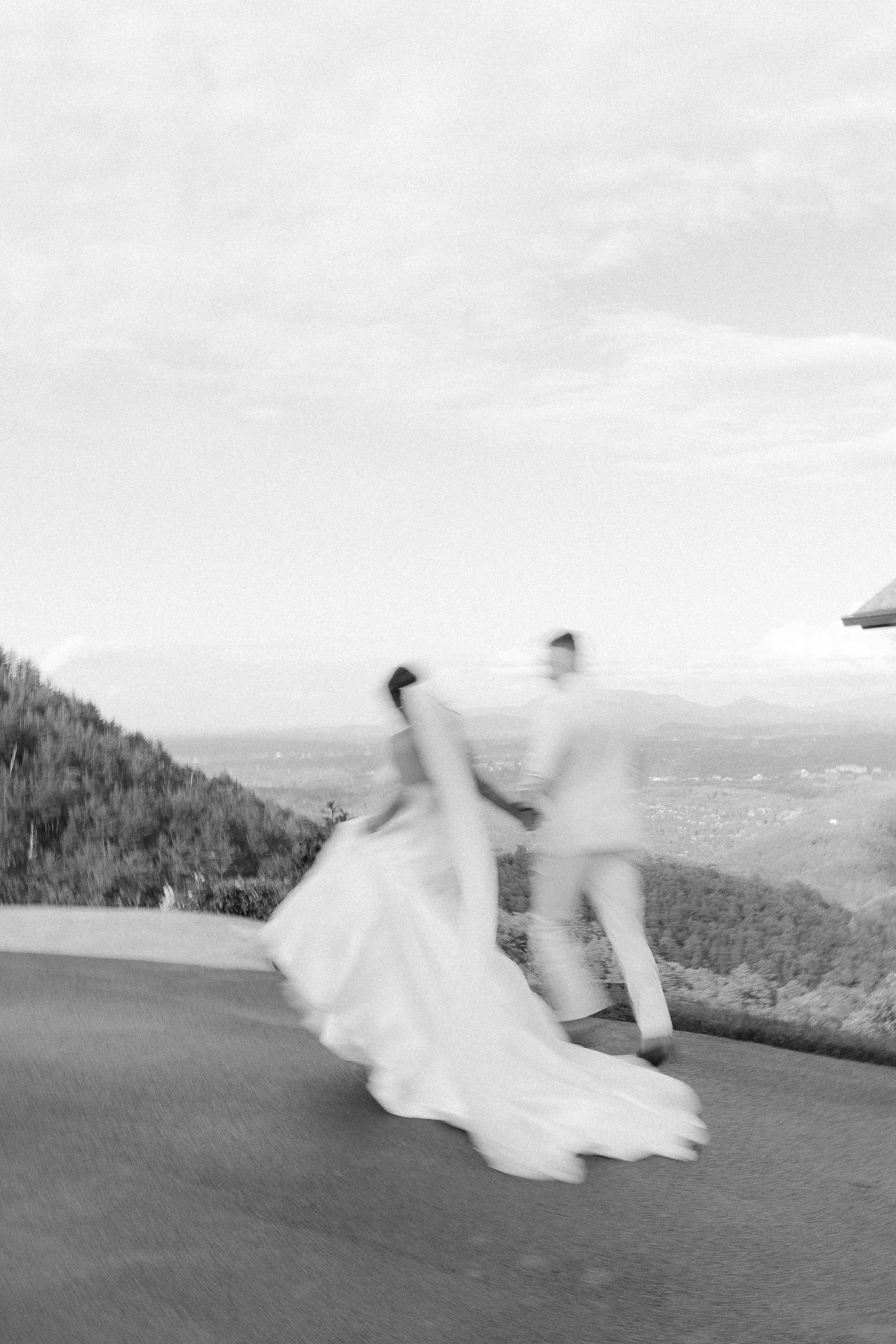 A couple embraces on a grassy mountain ridge with rolling blue mountains and valleys stretching into the distance.