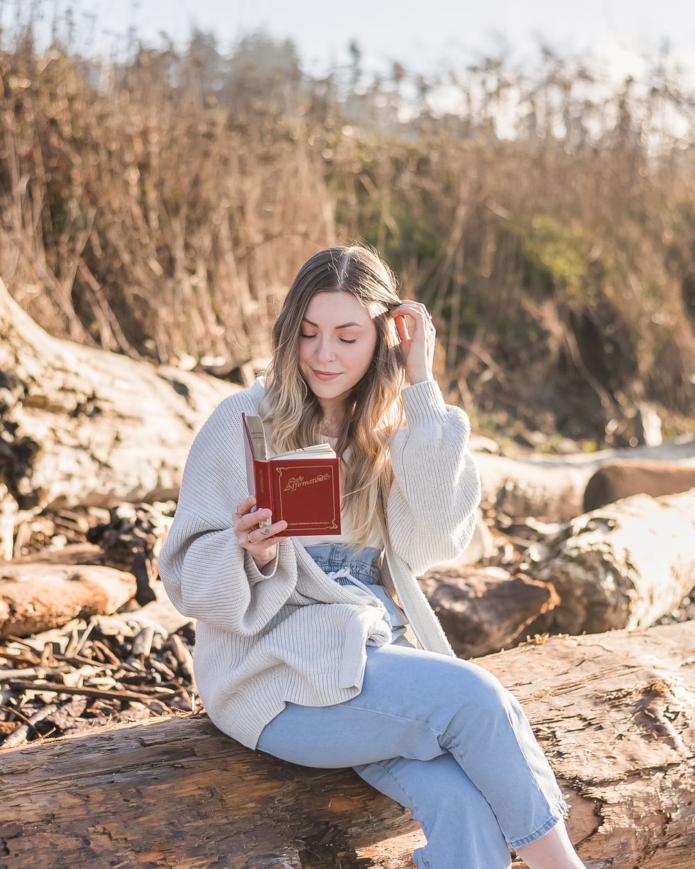 On a sunny spring morning a woman dressed in jeans and a sweater reads her book at the beach. She fixes her hair with her hand.