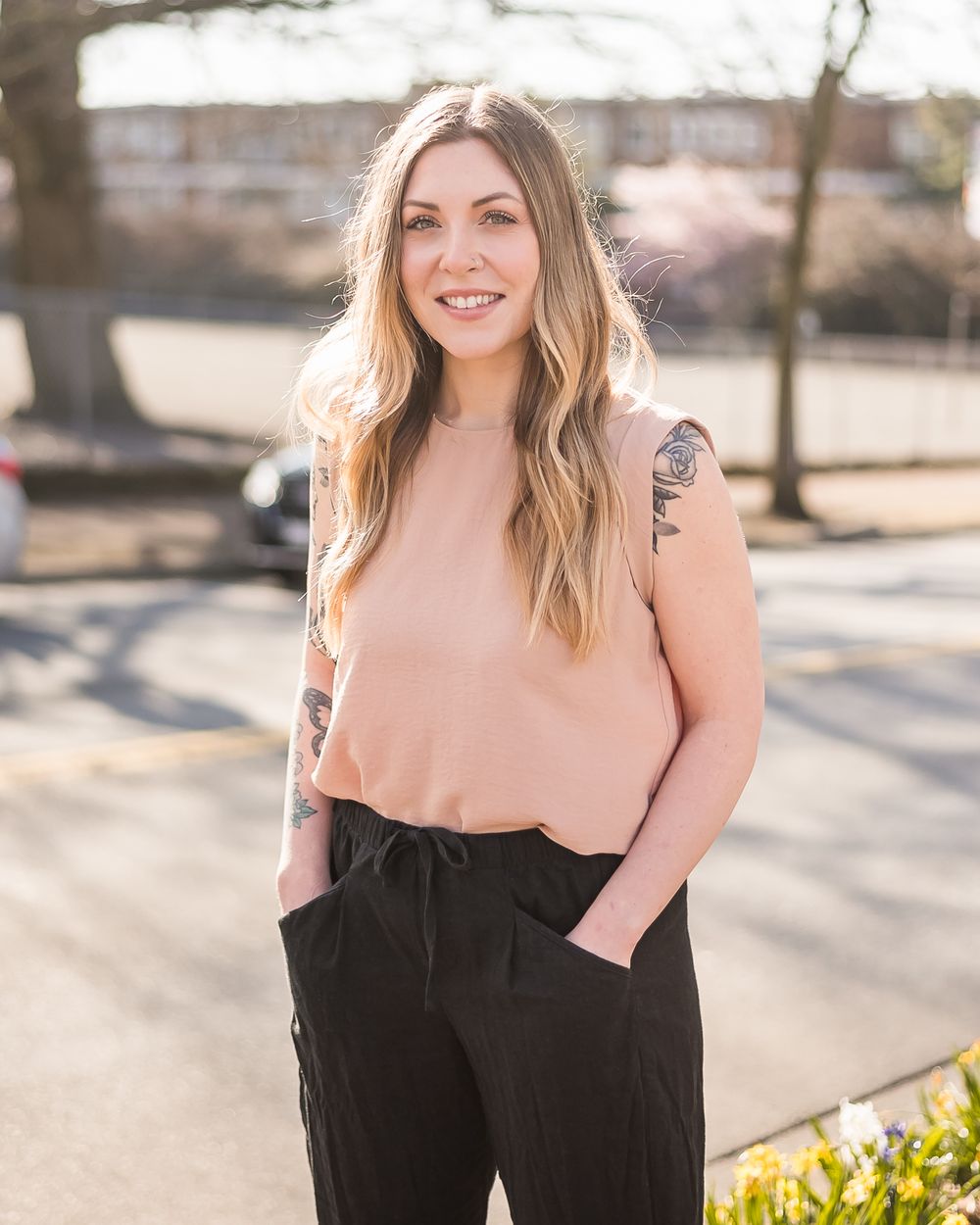 Standing in the bright sunshine a woman poses for a branding photo session in a peach shirt and black pants.