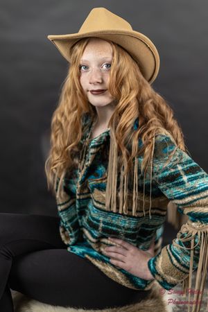 A portrait in western style clothing with a tan cowboy hat and teal fringed sweater against a dark background.