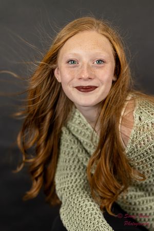 Portrait against dark backdrop showing windswept red hair, dark lipstick and sage green knit sweater.