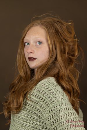 A portrait of someone with long wavy auburn hair wearing a sage green knit sweater against a dark brown background.