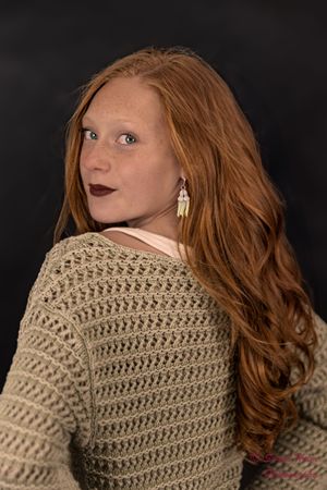 Portrait against dark background features long red hair and knit sweater with dramatic over-the-shoulder pose.