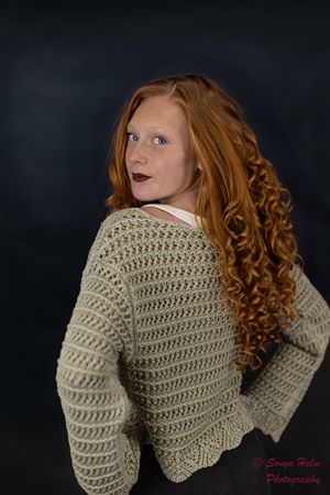 A long-haired figure wearing a cream knit sweater poses against a dark background in a studio portrait.