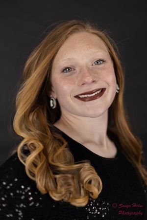 A portrait of someone with wavy strawberry blonde hair wearing a black top with dark lipstick against a dark background.