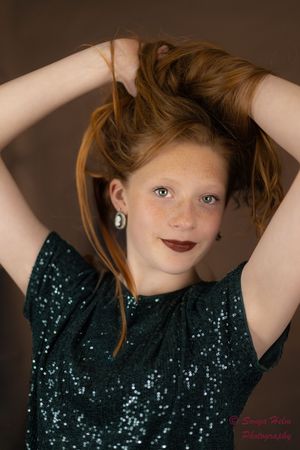 A person in a sparkly emerald green sequin top poses with their hands up, styling their ginger hair against a dark background.
