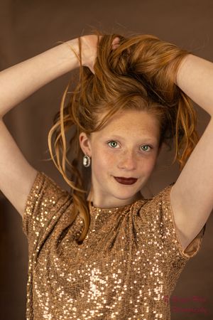 Young person in a sparkly gold sequin dress holding their red hair up while posing against a brown backdrop.