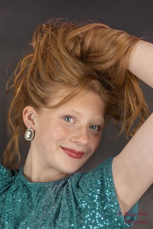 person in a sparkly turquoise sequin top with ginger hair and silver earrings poses against dark background.