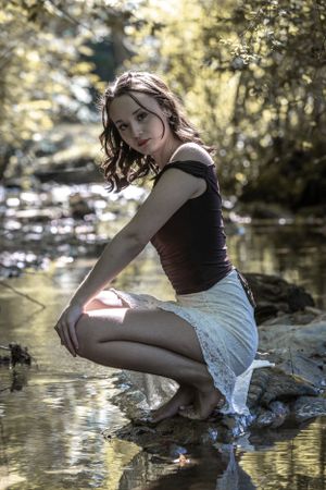 Artistic portrait of a person in black top by creek water with sunlit forest background.